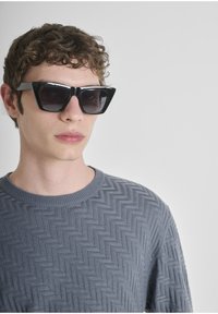 Occhiali da sole neri e angolari con lenti scure, abbinati a un maglione grigio texturizzato con motivo a spina di pesce e scollo rotondo.