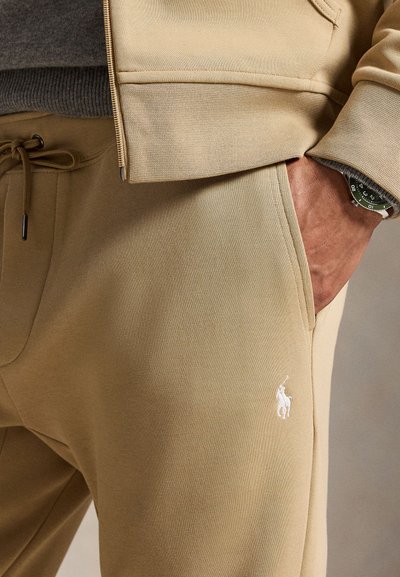 Gros plan d'une personne portant un pantalon de survêtement beige avec un logo cheval blanc brodé et une veste zippée assortie, main dans la poche, montre-bracelet visible.