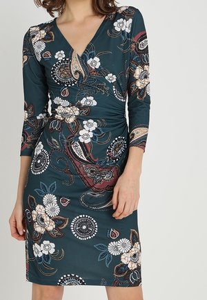 Vrouw draagt een donker teal knielange jurk met witte, beige en bordeaux bloem- en paisley patronen, V-hals en driekwart mouwen.