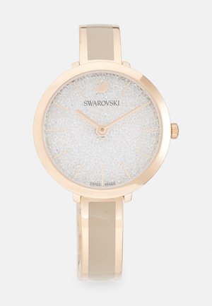Montre Swarovski avec un boîtier rond en ton or rose, un cadran texturé blanc et un bracelet brillant beige. Présente des marqueurs d'heure minimalistes et un logo.