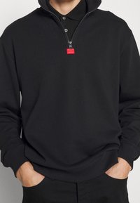 Sort sweatshirt med halvlynede krave, der har et lille rødt logomærke og elastiske ærmegab. Blødt, tekstureret stof med en afslappet pasform.