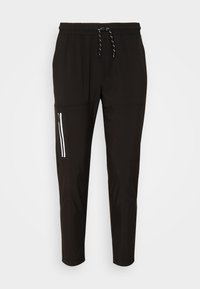 Svarta tapered byxor med elastisk midja, dragsko, och ficka med dragkedja med reflektortape på vänster lår mot vit bakgrund.