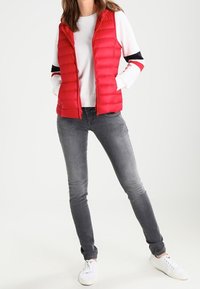 Röd pufferväst med vertikal quiltad textur, kombinerad med en vit långärmad skjorta med svarta och röda accenter, samt grå tighta jeans.