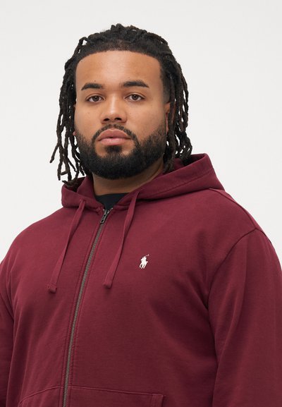 Homem com barba e dreadlocks, vestindo um hoodie com fecho éclair de cor vinho e um logótipo em branco de polo, de pé contra um fundo claro e simples.