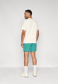 Μπλούζα κοντομάνικη εκρού, άνετη εφαρμογή, συνδυασμένη με σορτς σε τυρκουάζ. Λευκά αθλητικά παπούτσια και κάλτσες μέχρι τον αστράγαλο συμπληρώνουν το ντύσιμο. Ουδέτερο φόντο.