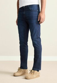 Dark blue denim jeans with a slim fit, featuring a classic five-pocket design and subtle contrast stitching. Worn with beige casual shoes.