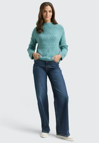 Ein tealfarbener Strickpullover mit rundem Ausschnitt und langen Ärmeln, kombiniert mit locker sitzenden dunkelblauen Jeans und weißen Turnschuhen.