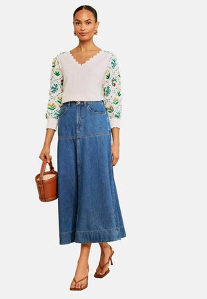 Kvinde iført en hvid strikket top med blomsterbroderede ærmer, en høj taljet blå denim midinederdel, brune sandaler med hæl og holder en brun bøtteformet taske.