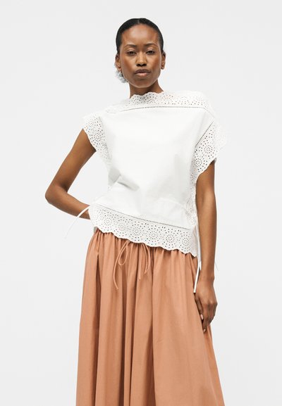 Γυναίκα που φοράει λευκό αμάνικο μπλουζάκι με δαντέλα eyelet και φούστα υψηλής μέσης, ανοιχτό καφέ, μακριά, μπροστά σε απλό φόντο.