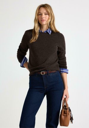 Kvinde med langt blond hår har en mørkebrun sweater over en blå stribet skjorte, mørke jeans og holder en brun håndtaske i højre hånd.