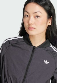 Čierna bomber bunda s bielymi prúžkami pozdĺž ramien, s celým zipsom a malým logom na hrudníku. Ľahký a odolný materiál.