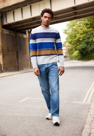 Jauns vīrietis staigā pa ielu zem tilta, valkājot pelēka, zila un brūna svītraina džemperi, zilas džinsa bikses un baltus sporta apavus.