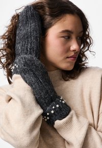 Donna con capelli ricci che indossa un maglione beige e muffole grigio scuro decorate con piccole perle bianche, che tiene le mani vicino al viso.