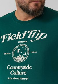 Groene katoenen t-shirt met ronde hals, voorzien van witte opdruk en een cirkelvormige afbeelding van bomen en vliegende vogels.