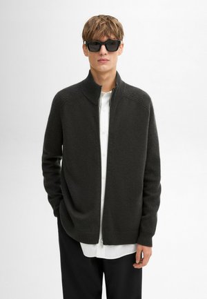 Cardigan grigio scuro a maglia con zip, texture a coste, colletto alto e maniche lunghe, indossato sopra una camicia bianca con bottoni. Occhiali da sole neri.