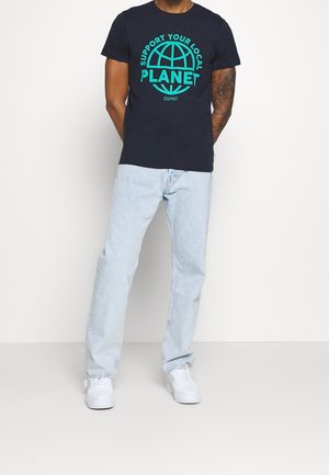 Mand iført en sort t-shirt med turkis tekst "Støt din lokale planet" og globusgrafik, lyseblå jeans og hvide sneakers.