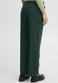 Wide-leg trousers in dark green fabric, featuring two back pockets and a smooth texture. Paired with black platform shoes.