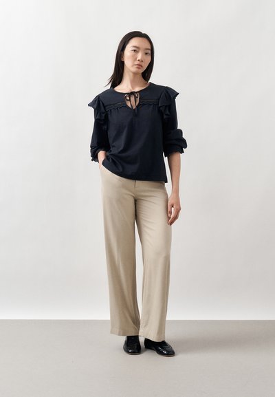 Femme portant un chemisier noir à manches longues volantées avec un col à nouer, un pantalon beige à jambes larges et des chaussures noires, debout devant un fond uni.