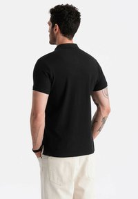 Černé polo tričko s klasickým límečkem a krátkými rukávy, nošené s béžovými kalhotami. Disponuje hladkou texturou a přiléhavým střihem.