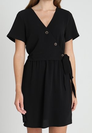 Femme portant une robe portefeuille courte noire avec trois boutons et un lien latéral, dotée de manches courtes et d'un décolleté en V.