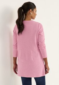 Long pink cardigan in a ribbed texture with a rounded neckline, long sleeves, and pockets. The hemline falls above the hips.