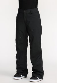 Pantalons de snowboard noirs à coupe ample, fabriqués en tissu durable. Comprend plusieurs poches et des coutures renforcées. Associés à des baskets noires.