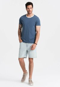 Niebieska bawełniana koszulka z białym kołnierzykiem, w zestawie z jasnymi dżinsowymi szortami. Model nosi beżowe sneakersy. Minimalistyczny design, casualowy krój.