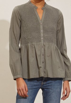 Mujer que lleva una blusa gris de manga larga con parte superior fruncida, botones delanteros y dobladillo tipo peplum fruncido, combinada con vaqueros azules.