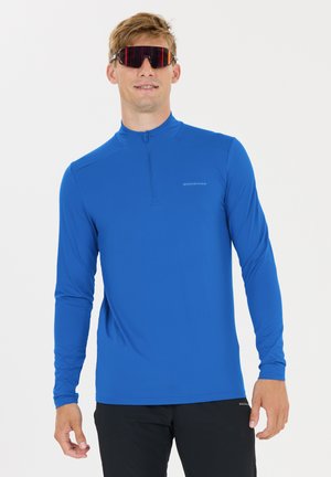 Man die een blauw atletisch shirt met lange mouwen en een halve ritssluiting draagt, zwarte broek en grote sportzonnebrillen met omhullend montuur.