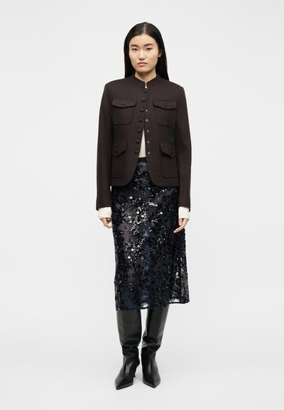 Γυναίκα που φοράει σκούρο καφέ σακάκι με κουμπιά, λευκό μακρυμάνικο τοπ, μαύρη midi φούστα με παγιέτες και μαύρες μπότες μέχρι το γόνατο, στέκεται σε λευκό φόντο.