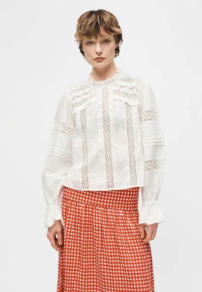 Γυναίκα που φοράει λευκό μπλουζάκι με δαντέλα στα άκρα, μακριά φουσκωτά μανίκια και ψηλό γιακά, σε συνδυασμό με κόκκινη και λευκή καρό φούστα.