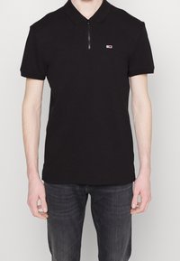 Svart polo skjorta med dragkedja i kragen, korta ärmar och en liten logotyp i röd och vit på vänster bröst. Strukturerad tyg.