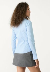 Blusa a righe blu chiaro con maniche lunghe, vestibilità sartoriale e orlo curvo, abbinata a una mini gonna grigia, vista da dietro.