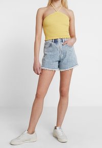 Žena ve žlutém halter topu, světlých modrých roztrhaných džínech a bílých teniskách, stojící s jednou rukou v kapse.