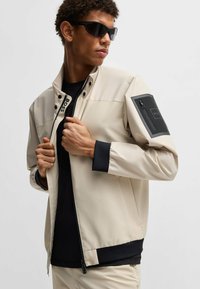 Giacca beige con un design strutturato, caratterizzata da una chiusura a zip, dettagli neri sulle maniche e un logo sul petto e sulla tasca.
