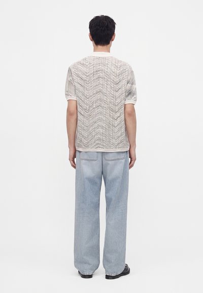 Homme aux cheveux courts et foncés, vu de dos, portant une chemise blanche texturée à manches courtes, un jean large bleu clair et des chaussures noires.