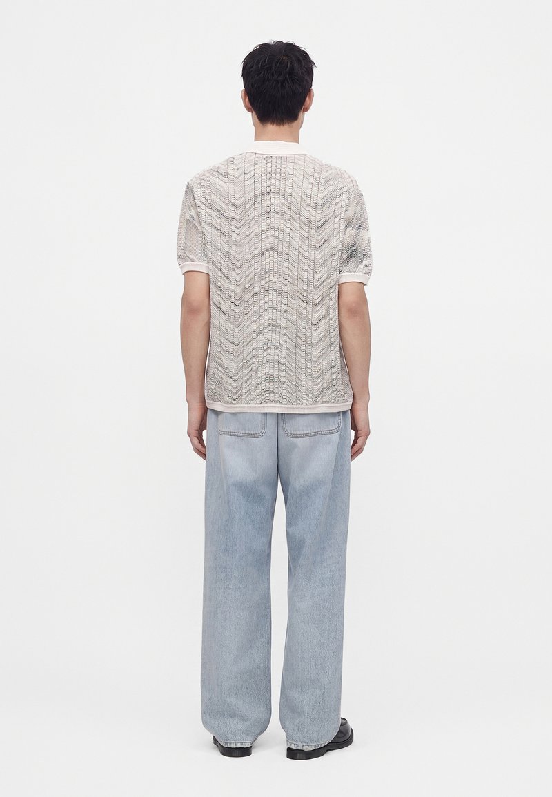 Hombre con cabello corto y oscuro de espaldas, lleva una camisa blanca texturizada de manga corta, jeans anchos de color azul claro y zapatos negros.