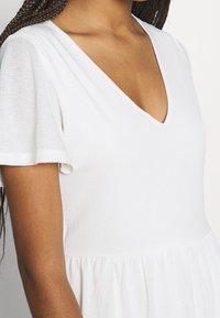 Bílé šaty s krátkým rukávem a výstřihem do V, s texturovanou látkou a řaseným pasem, vytvářející rozšířenou siluetu.