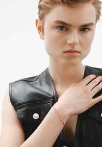 Top sin mangas de cuero negro con un destacado detalle de herrajes plateados, acompañado de una delicada pulsera de infinito dorada en la muñeca.