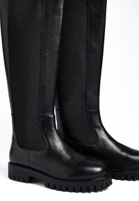 Črne škornje do kolen, izdelane iz gladke usnje, z teksturirano elastično ploščo, ki ima čvrst podplat in zaobljene prste.
