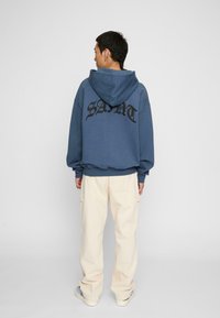 Felpa blu scuro con grande testo nero "SAINT" sulla schiena, abbinata a pantaloni cargo crema. Design casual, texture morbida e vestibilità rilassata.
