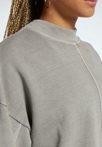 Ljusgrå sweatshirt med ribbad rund hals, sänkta axlar och en slät yta. Synliga sömmar som framhäver sömmarna.