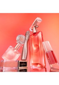 Vari cosmetici Lancôme tra cui profumo, mascara, ombretto e gloss per labbra disposti su una superficie riflettente con uno sfondo rosa-arancione.