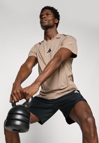 Kettlebell noir tenue à deux mains par une personne portant un t-shirt de sport beige avec des accents noirs et un short noir, sur un fond neutre.