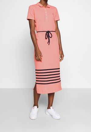 Osoba ubrana w koralowo-różową sukienkę polo z czarnym paskiem na sznurku, czarne poziome paski przy krawędzi i białe trampki, stojąca na tle gładkiej ściany.