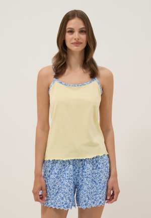 Young woman wearing light yellow camisole with blue floral trim and matching blue floral shorts, standing against plain background.