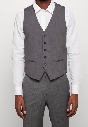 Mann trägt weißes Langarmhemd, graue geknöpfte Weste und passende graue Hose, steht vor einem einfarbigen Hintergrund.