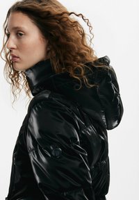 Giacca a vento nera con una texture lucida e liscia, dotata di cappuccio e tasche con zip. Il design trapuntato enfatizza l'isolamento e il calore.