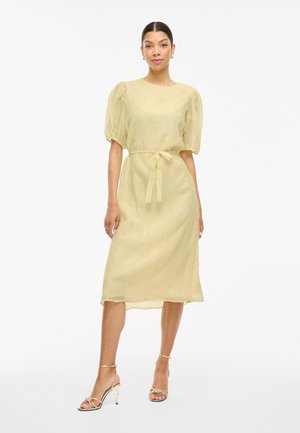 Woman wearing a light yellow midi dress with puff sleeves, a tied waist belt, and gold strappy high-heeled sandals standing against white background.