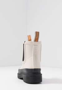 Botín de tobillo de goma beige con suela negra, paneles laterales elásticos y lengüetas en naranja y beige. Superficie texturizada y diseño minimalista.